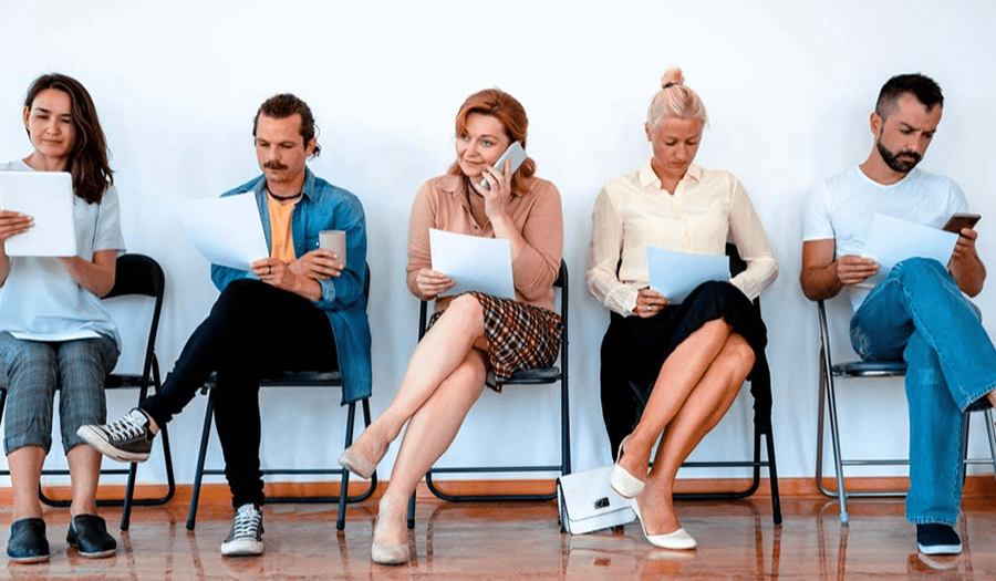 a group of people sit on chairs against a white wall, they all read and fill out paper work.