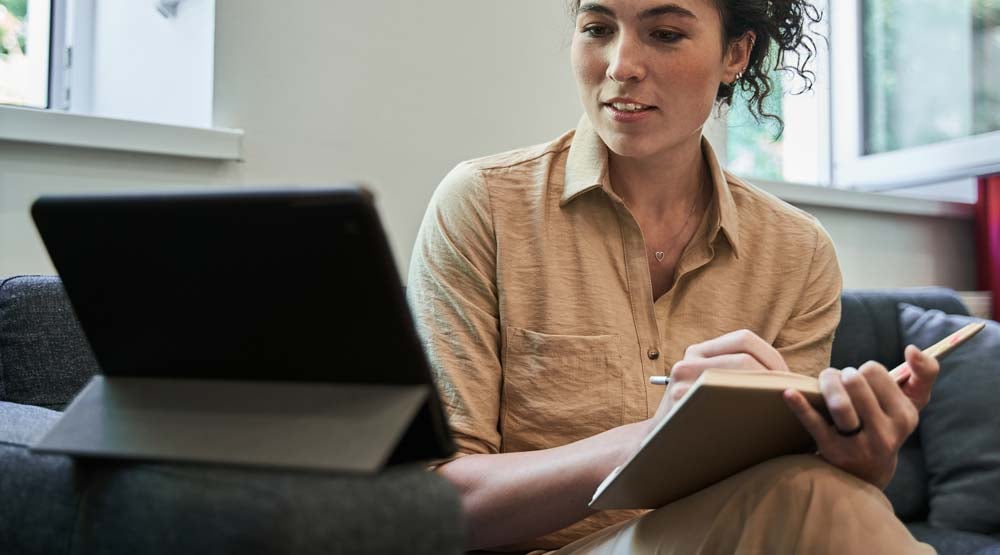 A young women at home takes notes while attending video meeting on her tablet