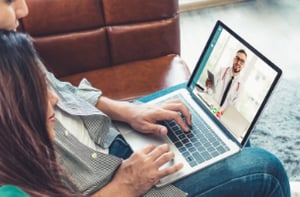 A young couple on a couch communicate with a doctor via their laptop.